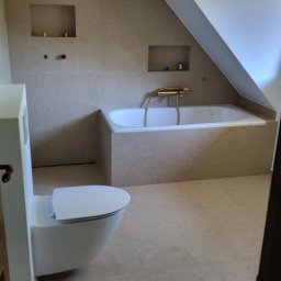 ANDBUD - Beżowa łazienka na poddaszu ze skośnym sufitem, z zabudowaną wanną z baterią w kolorze złotym, podwieszaną toaletą i wnękami w ścianie.