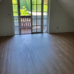 Poddasze z drewnianą podłogą i widokiem na balkon z zielenią w tle, surowe drewniane elementy konstrukcyjne na suficie.