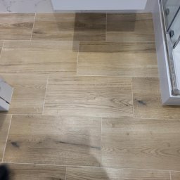 ANDBUD - Ułożona podłoga z płytek imitujących drewno w łazience, widoczne fragmenty szafki pod umywalką i kabiny prysznicowej, robocze buty na podłodze.