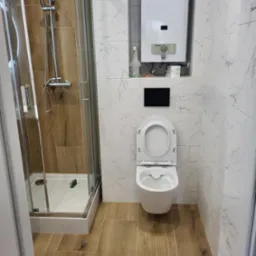 Nowoczesna łazienka z kabiną prysznicową ze srebrnymi profilami, podwieszaną toaletą i piecykiem gazowym na tle białych płytek imitujących marmur oraz brązowych płytek podłogowych imitujących drewno.