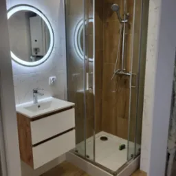 Nowoczesna łazienka z kabiną prysznicową w rogu, umywalką z szafką w odcieniach drewna i białego oraz okrągłym lustrem z podświetleniem LED. Na ścianach płytki imitujące marmur, a w kabinie...