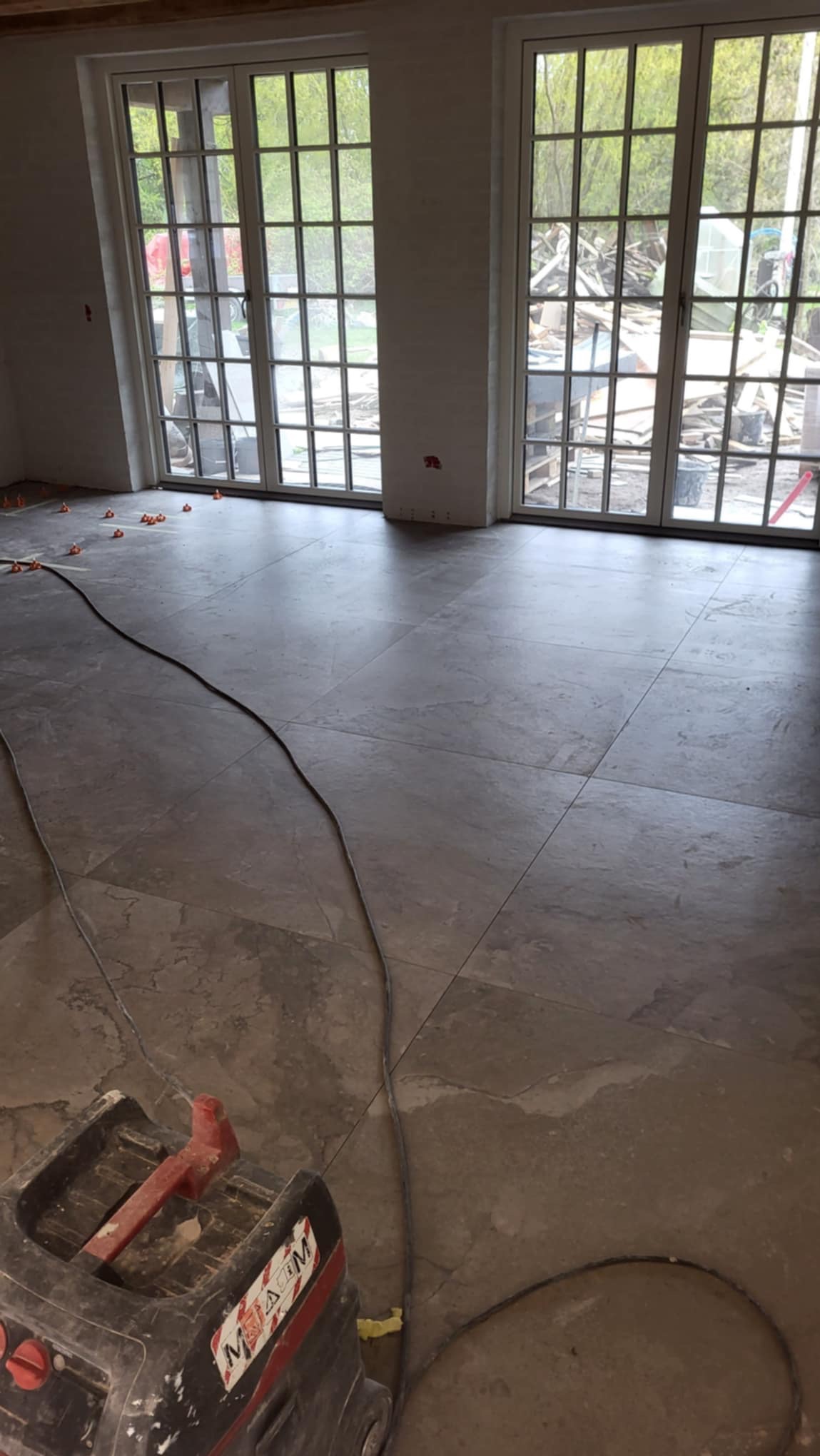 Układanie płytek podłogowych w jasnym pomieszczeniu z dużymi oknami, widoczne narzędzie do fugowania i pomarańczowe podkładki dystansowe.