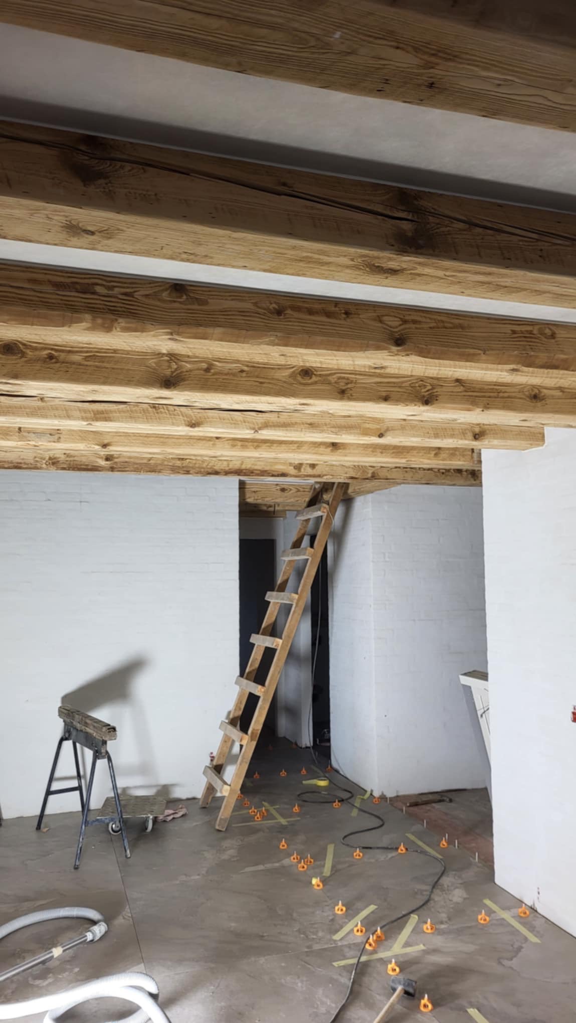Wnętrze w trakcie remontu z drewnianymi belkami stropowymi, drabina oparta o ścianę, poziomowanie podłogi za pomocą specjalnych klipsów i poziomicy laserowej.