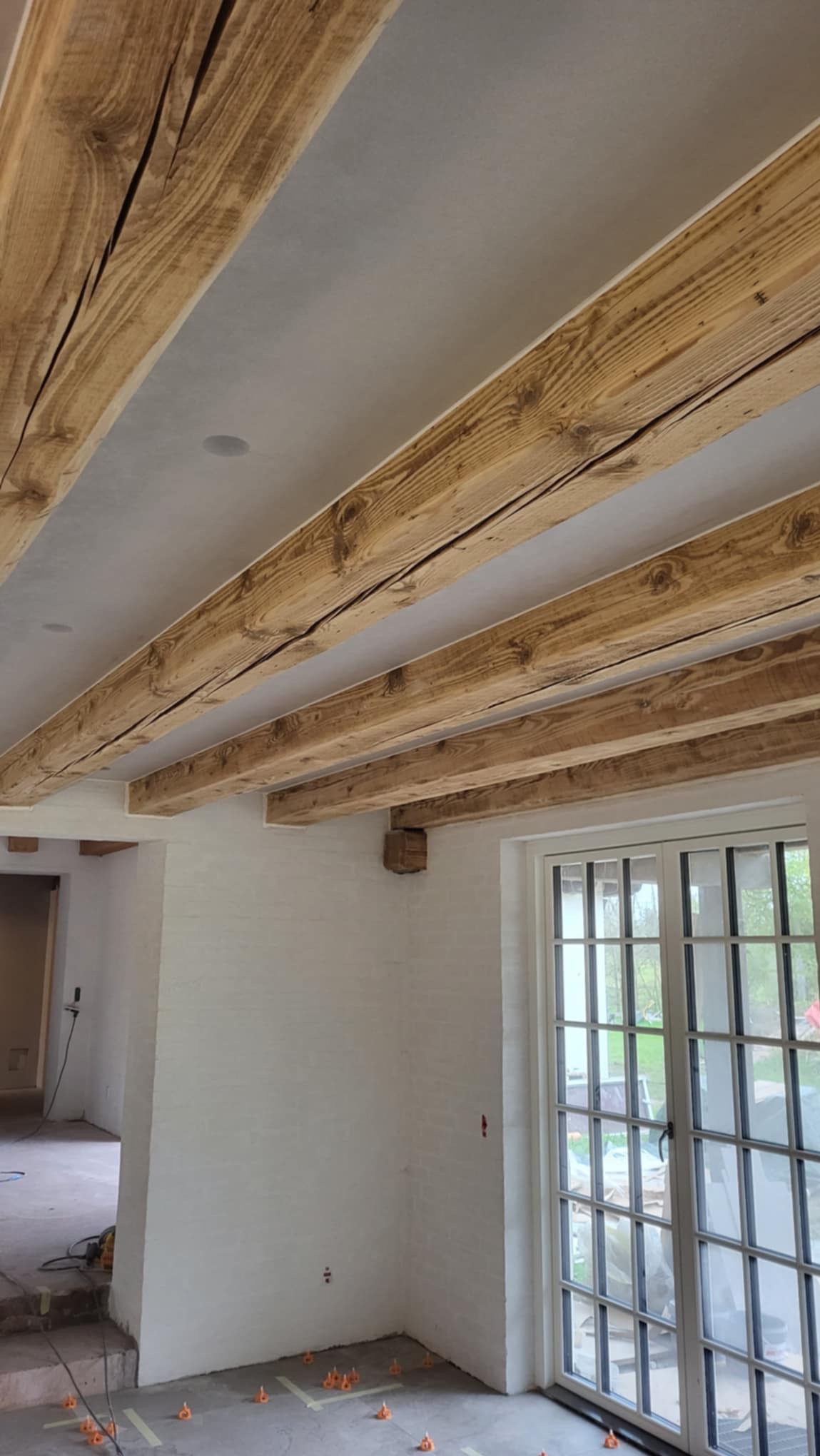 Wnętrze w trakcie prac wykończeniowych: sufit z widocznymi drewnianymi belkami, białe ściany z cegły, okno z białymi szprosami, na podłodze plastikowe krzyżyki dystansowe do układania płytek.