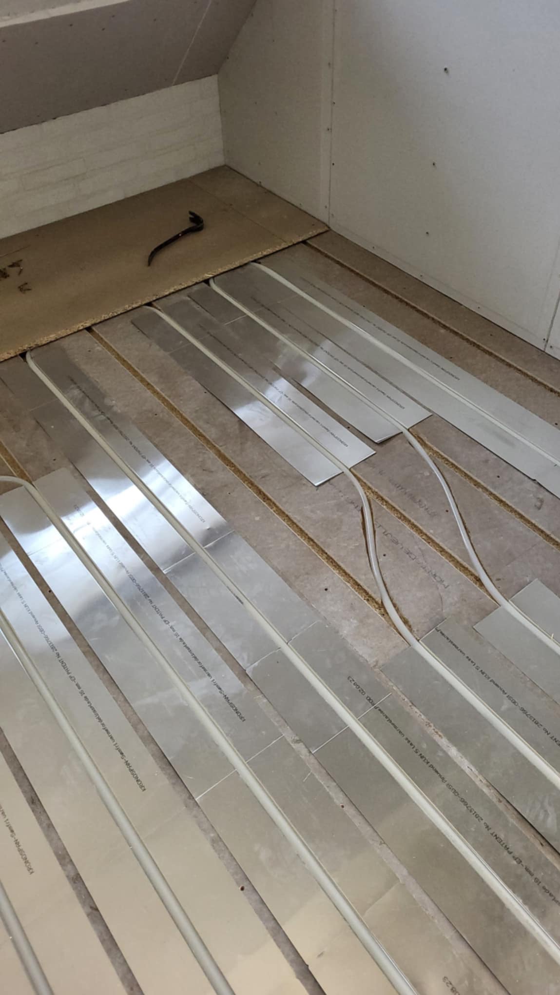 Instalacja ogrzewania podłogowego w poddaszu: widoczne płyty rozprowadzające ciepło z rurami, przygotowane do dalszych prac wykończeniowych, z narzędziem w rogu pomieszczenia.