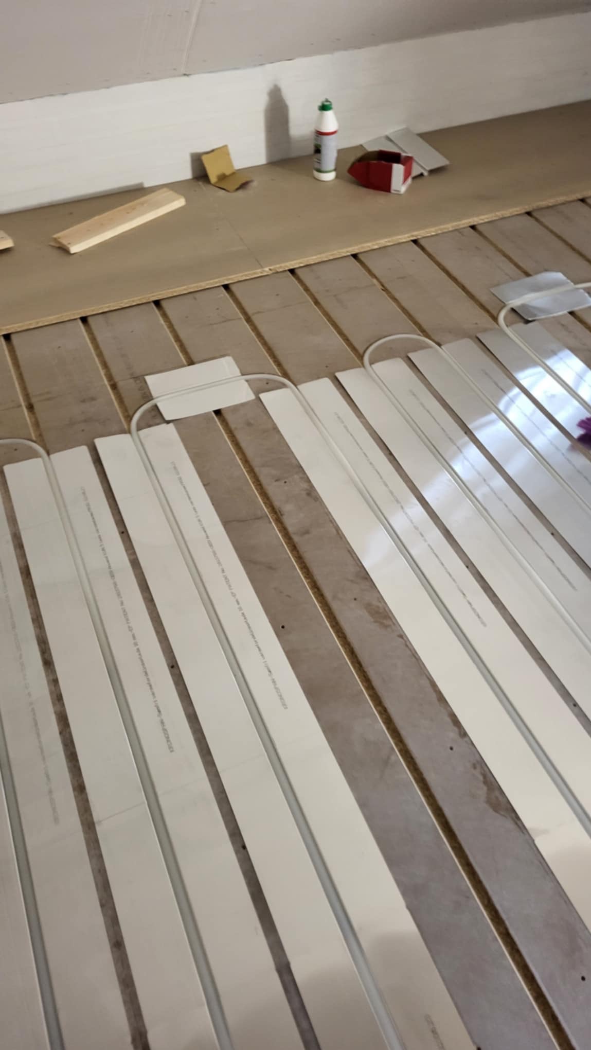 Instalacja ogrzewania podłogowego w systemie suchym, widoczne białe panele grzewcze ułożone na drewnianej podłodze, z akcesoriami montażowymi i klejem w tle.