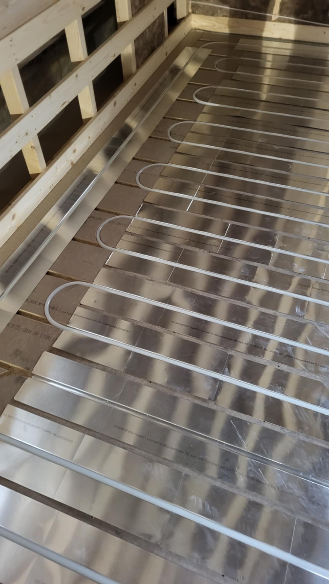 Montaż ogrzewania podłogowego: rury grzewcze ułożone na aluminiowych płytach rozpraszających ciepło, widoczne drewniane elementy konstrukcyjne poddasza.