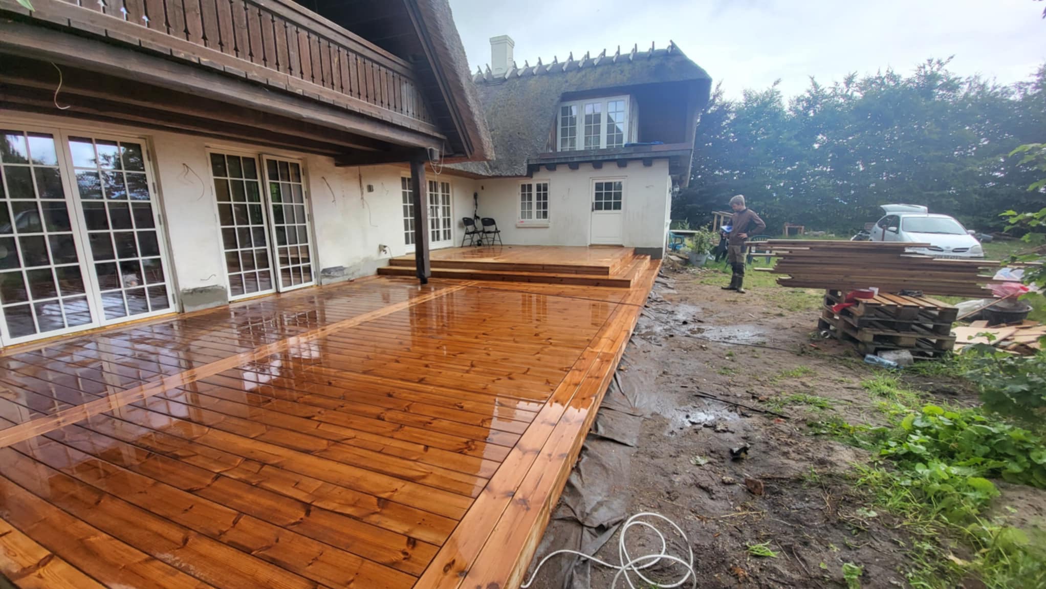 Świeżo zaimpregnowany, lśniący drewniany taras przy domu z krytym strzechą dachem w Górowie Iławeckim. W tle widoczne materiały budowlane i pracownik.