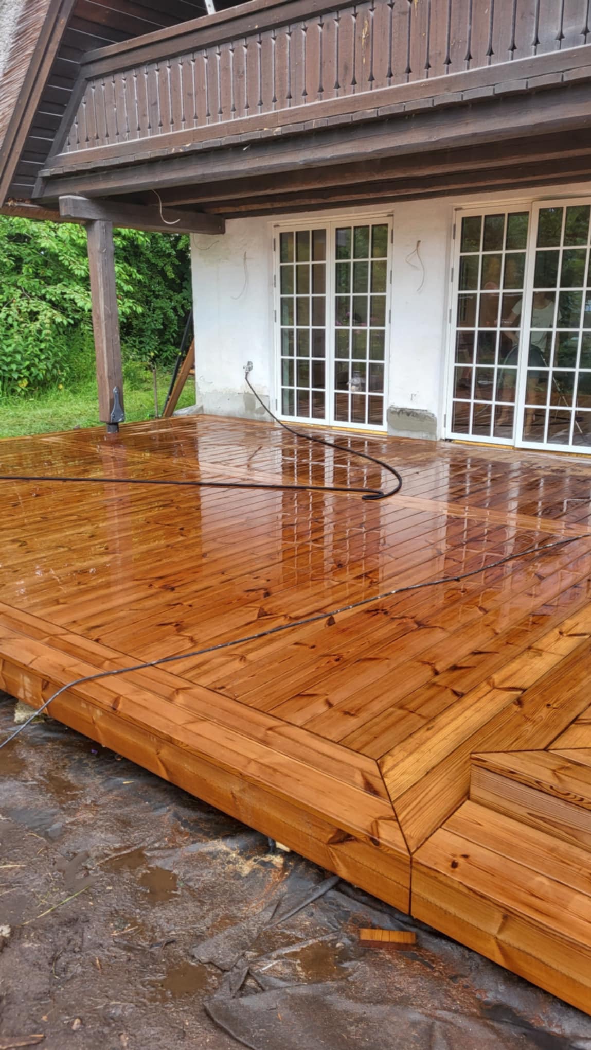 Świeżo zaimpregnowany, mokry drewniany taras z widocznymi śladami wody po myciu, z elementami dekoracyjnymi na obrzeżach, na tle białego domu z dwuskrzydłowymi oknami.