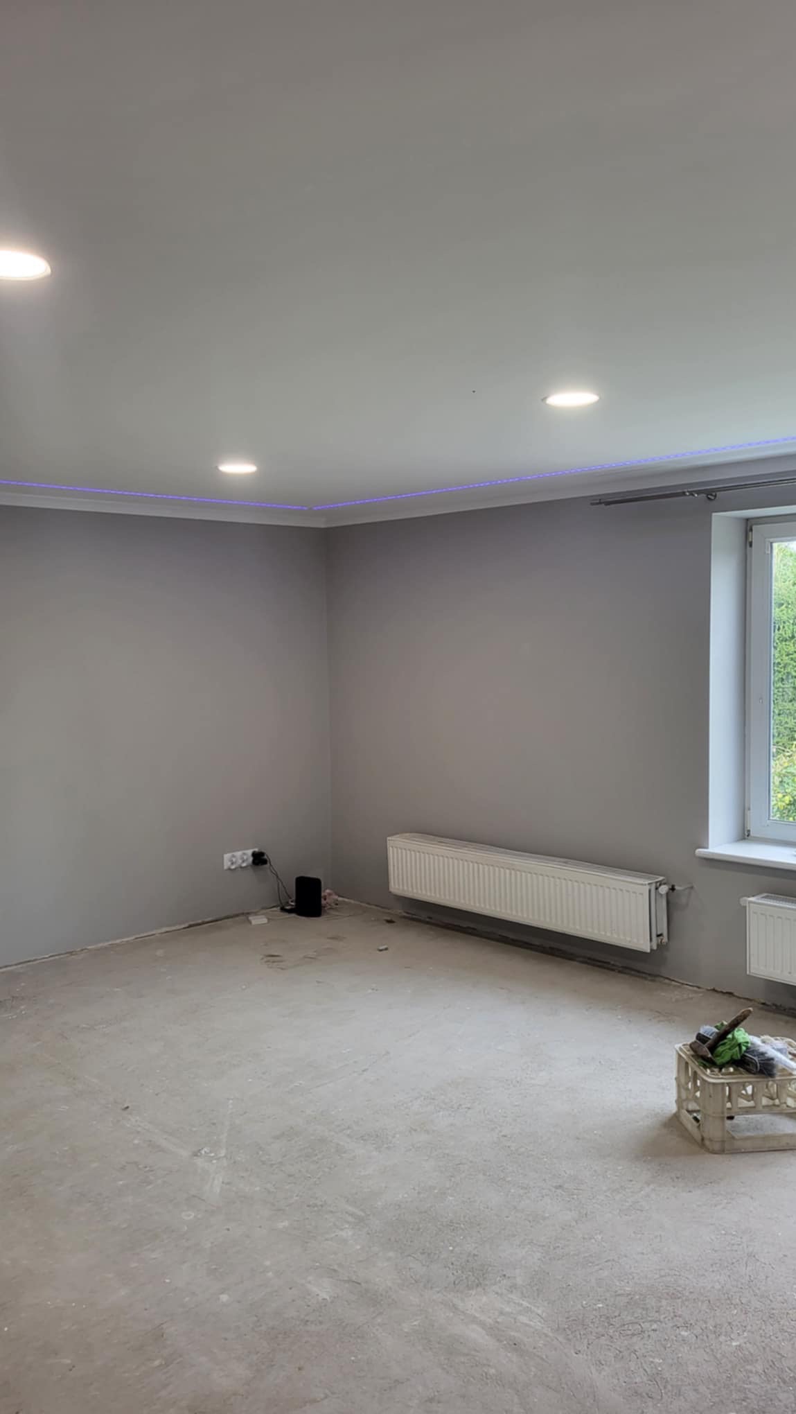 Szare, świeżo pomalowane wnętrze pokoju z grzejnikami, oświetleniem sufitowym i listwą LED pod sufitem. Podłoga w trakcie przygotowania pod wykończenie, widoczny plastikowy pojemnik z narzędziami.