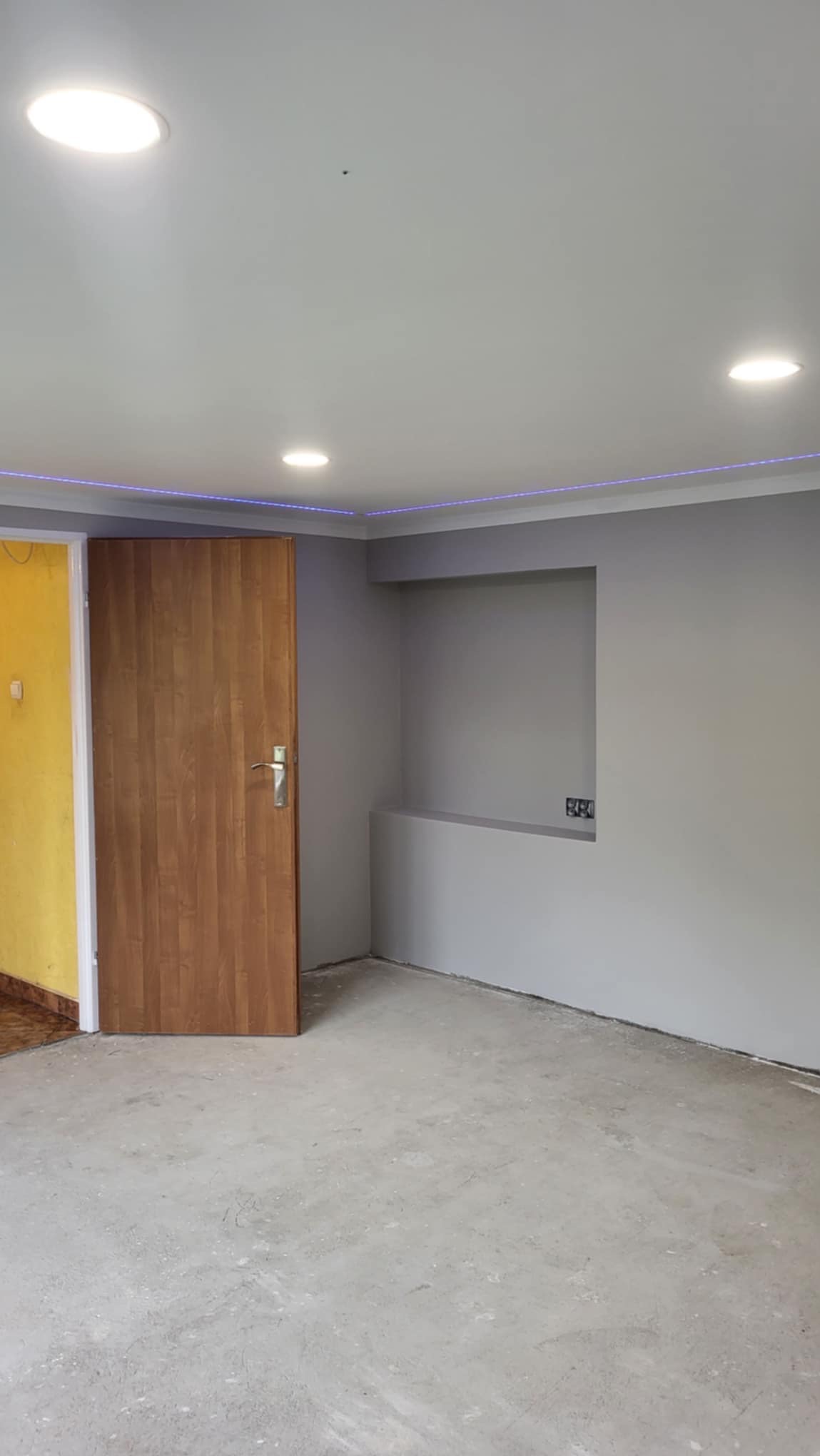 Szare wnętrze pokoju w trakcie prac wykończeniowych, z otwartymi drewnianymi drzwiami, widoczną instalacją elektryczną w ścianie i oświetleniem LED na suficie.