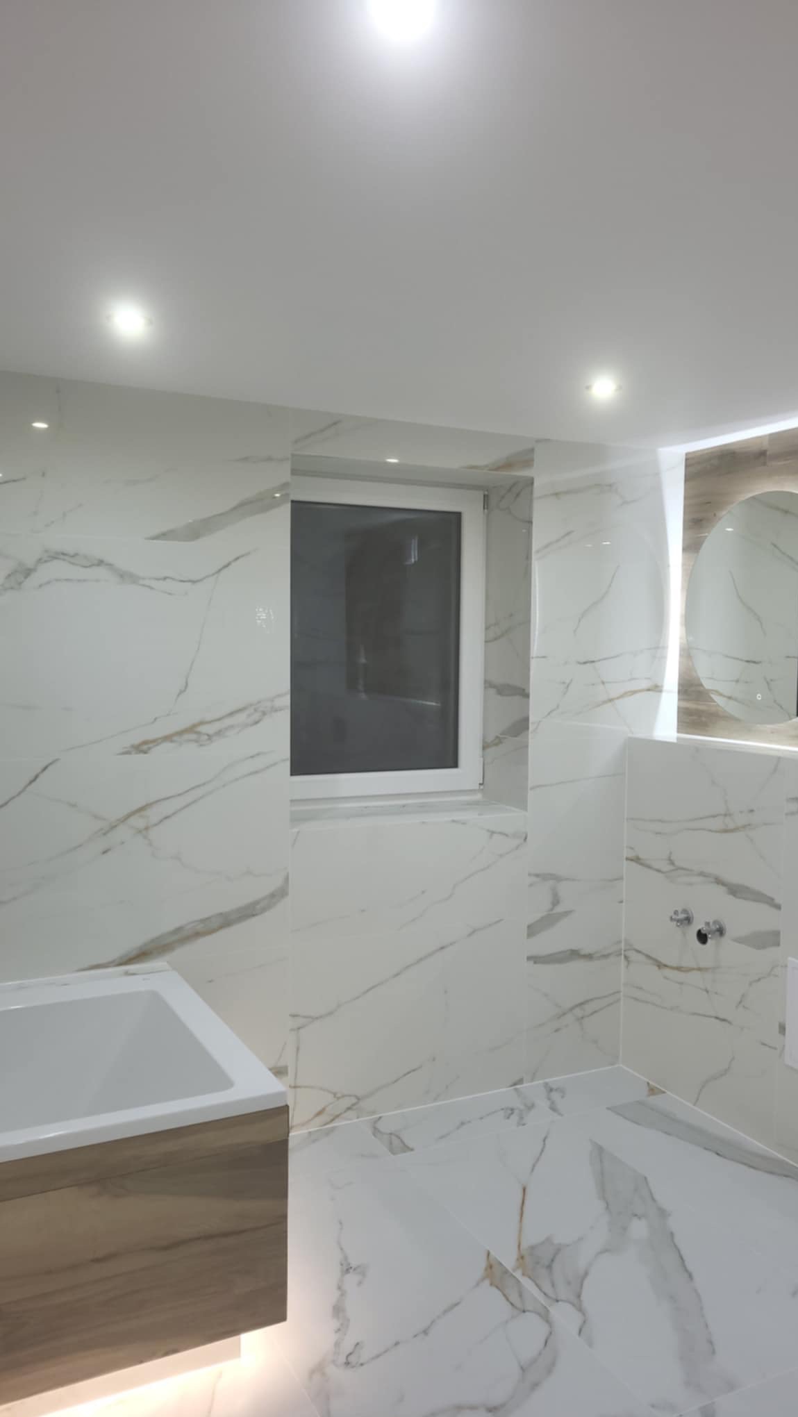 Nowoczesna łazienka wykończona białymi płytkami imitującymi marmur, z widoczną umywalką z drewnopodobną szafką, oknem i okrągłym lustrem z podświetleniem.