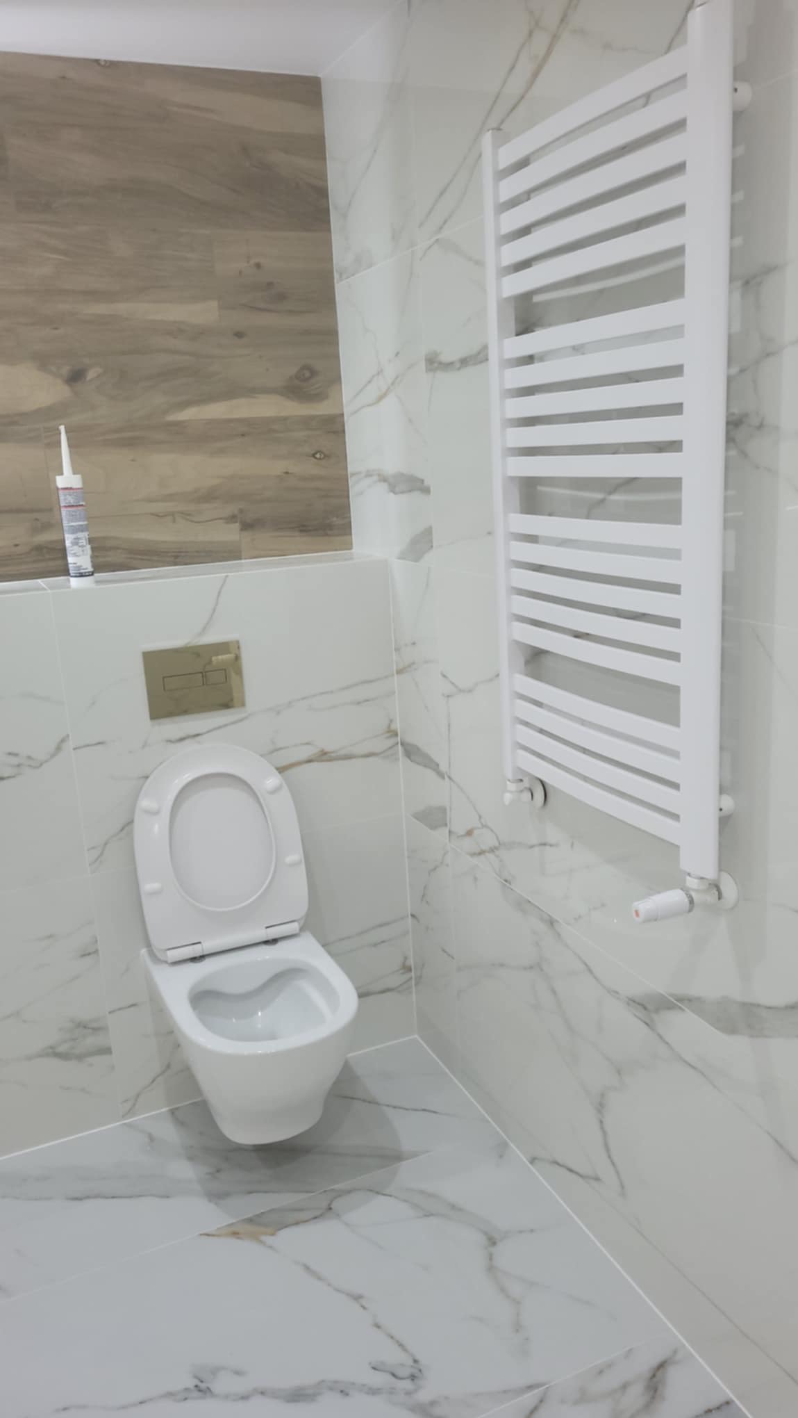 Wykończona łazienka z toaletą wiszącą, grzejnikiem drabinkowym i płytkami imitującymi marmur na ścianach i podłodze, z akcentem drewnopodobnych płytek na fragmencie ściany. Widoczna tuba silikonu...