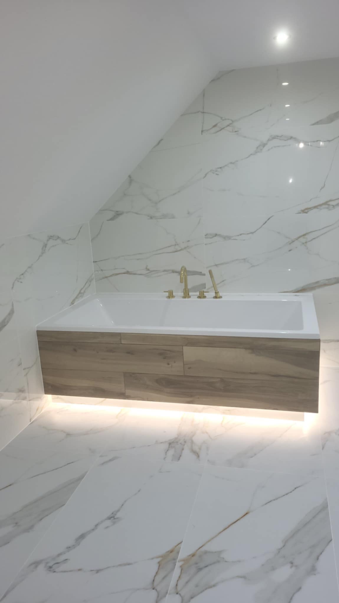 Nowoczesna łazienka w odcieniach bieli i beżu z wanną obudowaną drewnopodobnymi panelami i podświetleniem LED, marmurowe płytki na ścianach i podłodze, złota armatura.