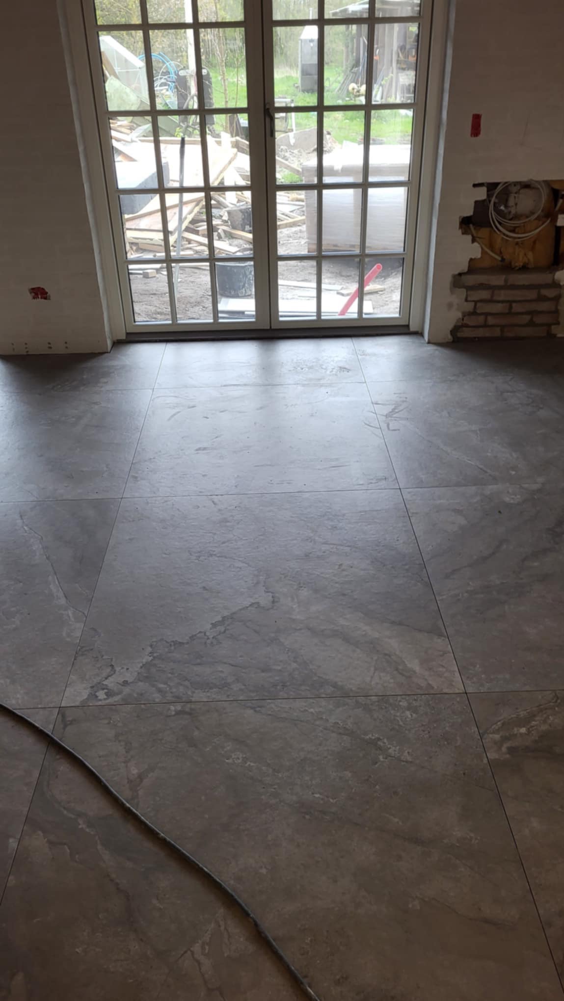 Szare, wielkoformatowe płytki podłogowe imitujące kamień, ułożone w pomieszczeniu z dużym oknem balkonowym i fragmentem niezabezpieczonej instalacji elektrycznej w ścianie.