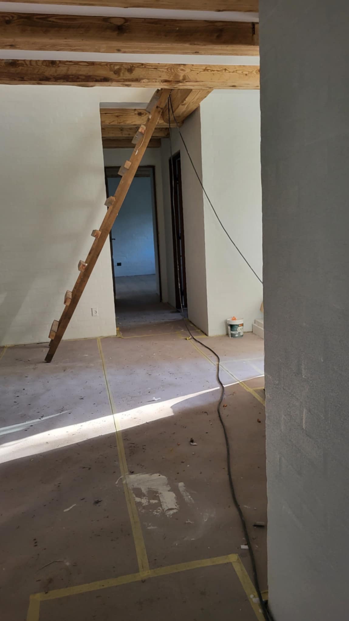 Wnętrze domu w trakcie prac wykończeniowych, widoczne drewniane belki stropowe, przystawiona drabina, ściany po gładziowaniu, podłoga zabezpieczona folią malarską.