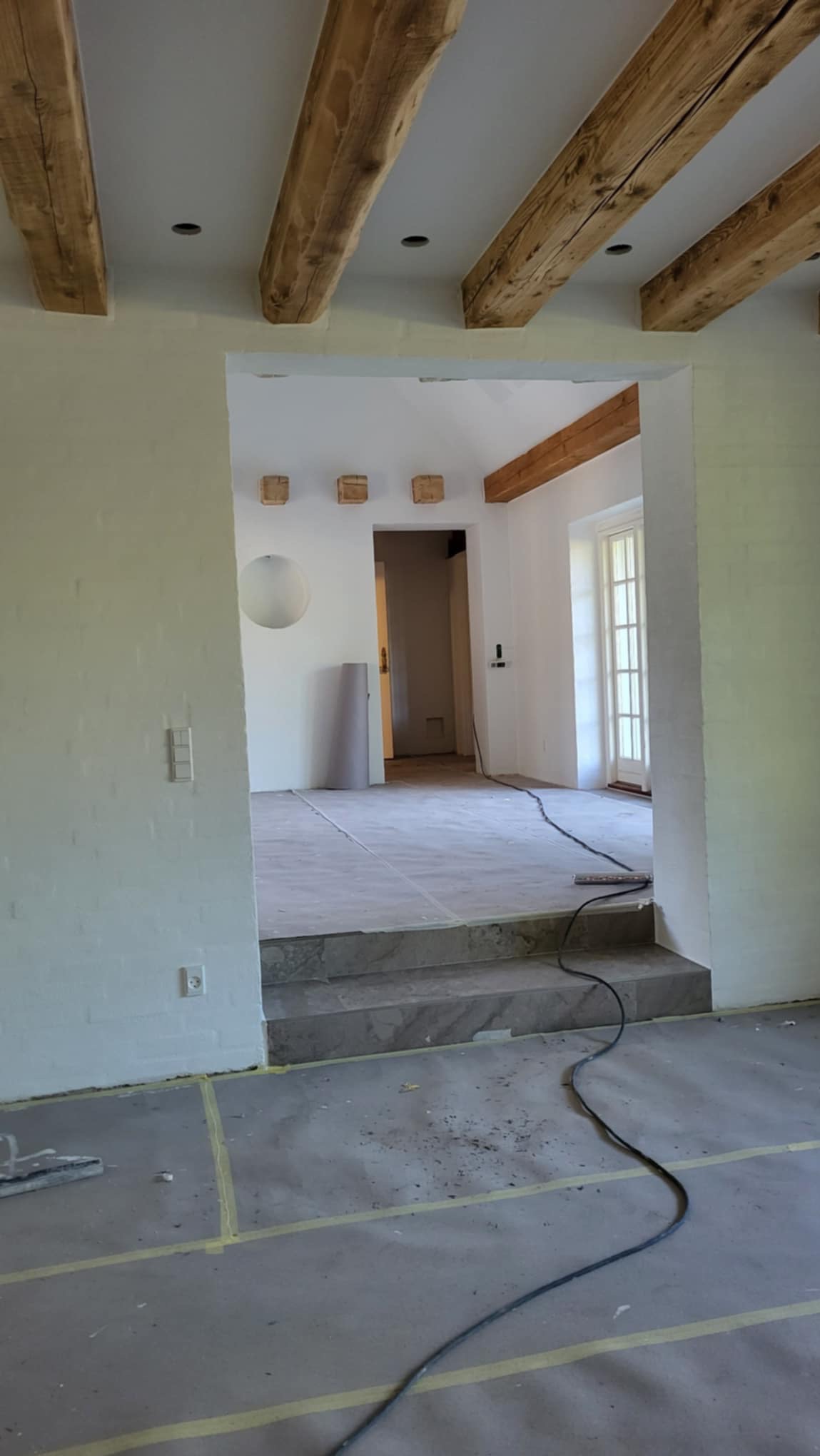 Wnętrze domu w trakcie remontu: widoczne surowe, drewniane belki stropowe, świeżo pomalowane ściany na biało, zabezpieczona podłoga folią i taśmą malarską, schody wyłożone marmurem.
