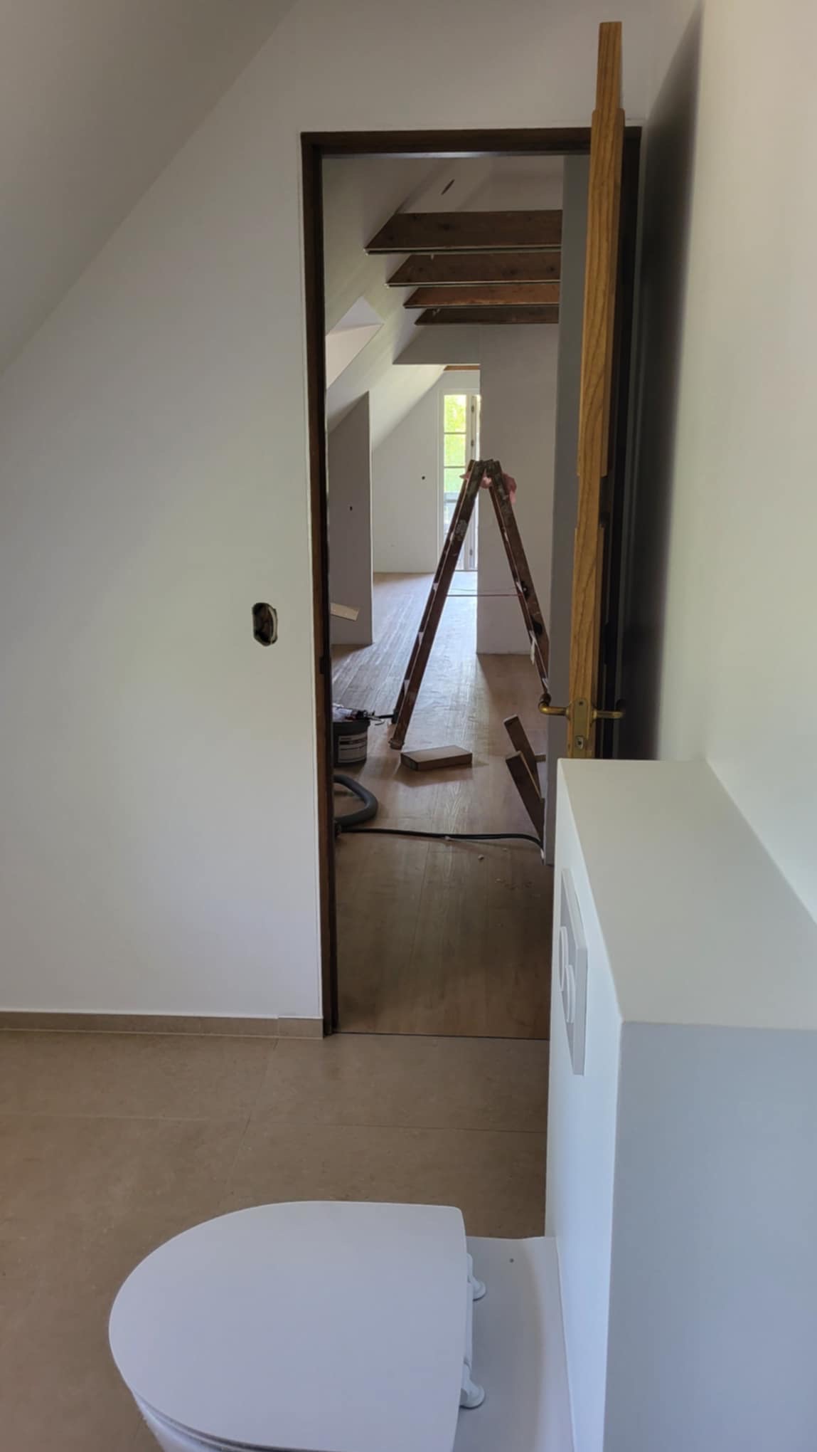 Wnętrze domu w trakcie remontu, widoczna drabina w przejściu, podłoga, ściany w jasnych kolorach i fragment toalety na pierwszym planie.