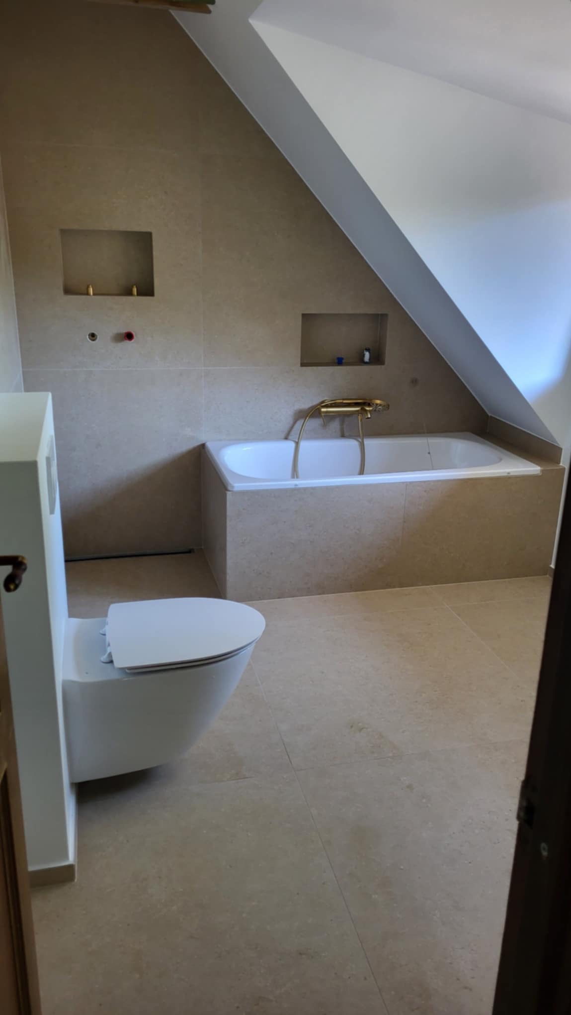 Beżowa łazienka na poddaszu ze skośnym sufitem, z zabudowaną wanną z baterią w kolorze złotym, podwieszaną toaletą i wnękami w ścianie.