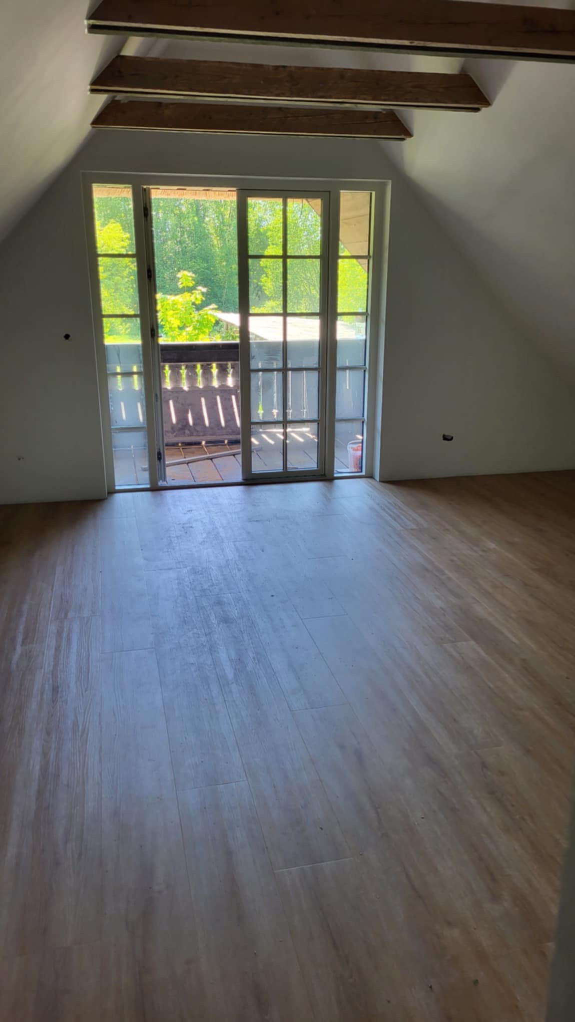 Poddasze z drewnianą podłogą i widokiem na balkon z zielenią w tle, surowe drewniane elementy konstrukcyjne na suficie.