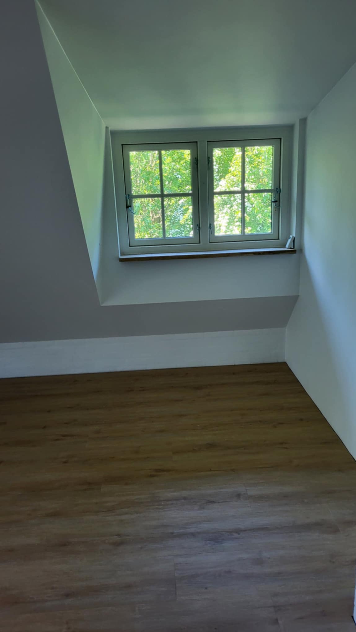 Poddasze po remoncie z drewnianą podłogą, białymi ścianami i oknem z widokiem na zieleń.