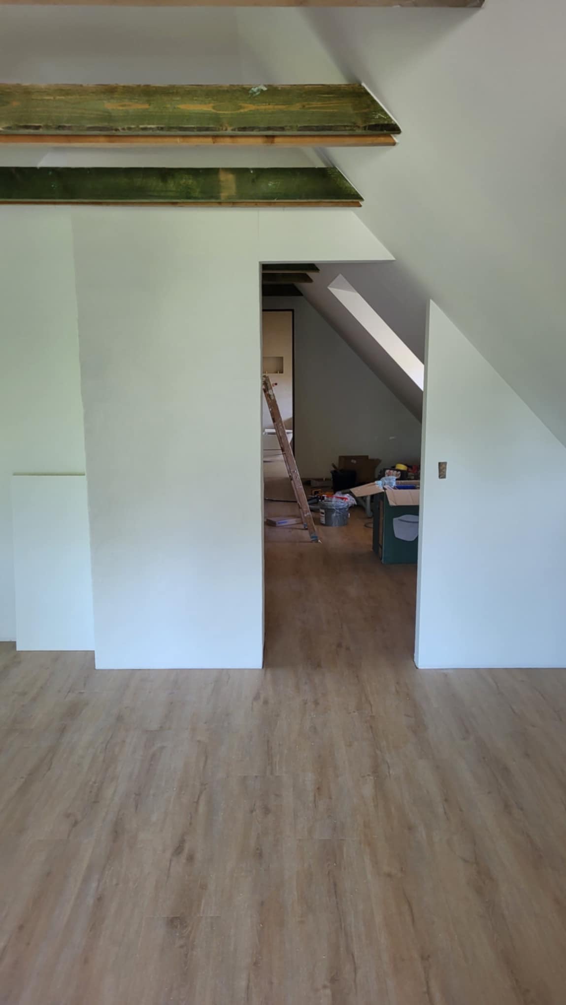 Poddasze w trakcie remontu, widoczne drewniane belki stropowe, ściany przygotowane do malowania, fragment podłogi z paneli, drabina i narzędzia w głębi pomieszczenia.