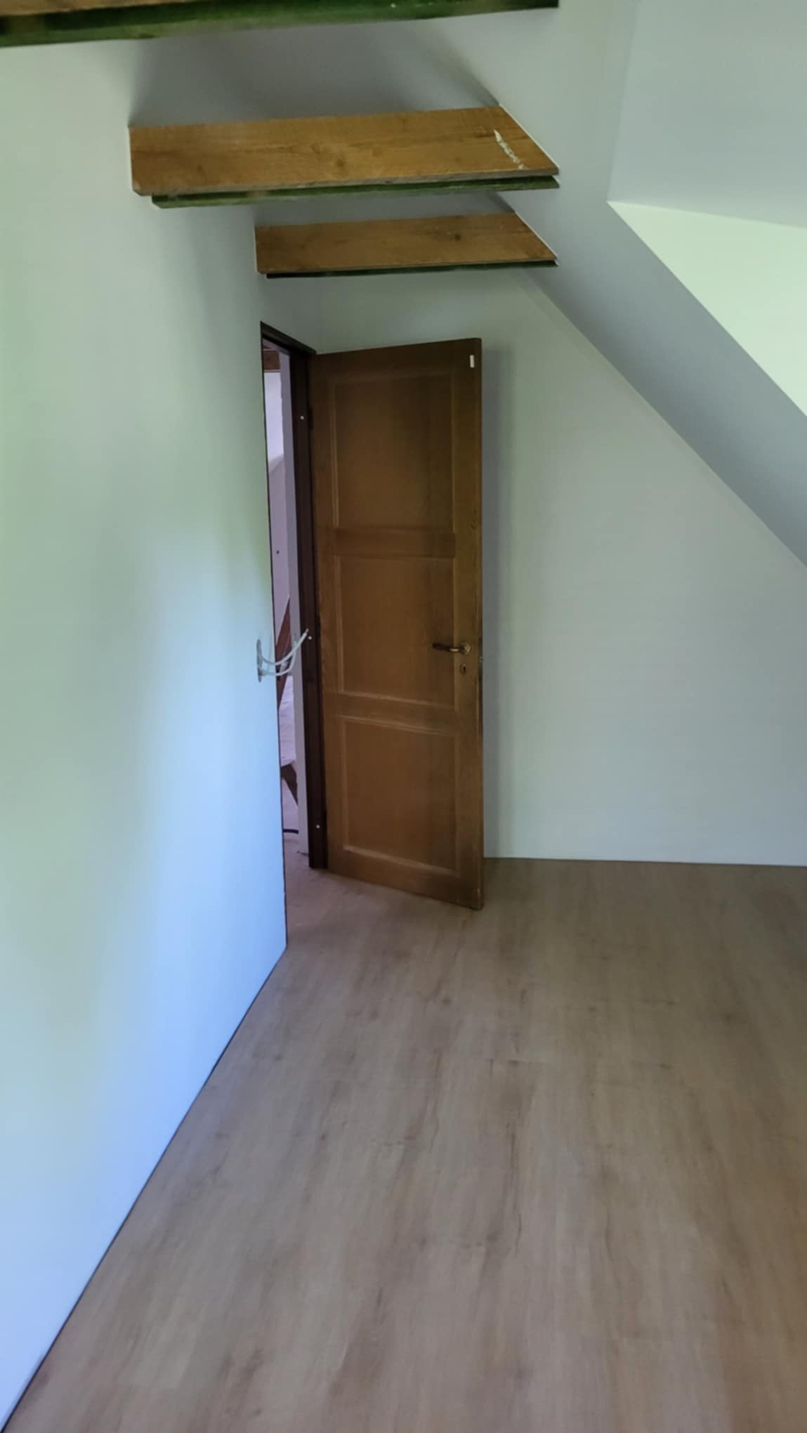 Poddasze po remoncie z drewnianą podłogą, otwartymi drewnianymi drzwiami i pomalowanymi na biało ścianami, widoczne drewniane elementy konstrukcyjne pod sufitem.