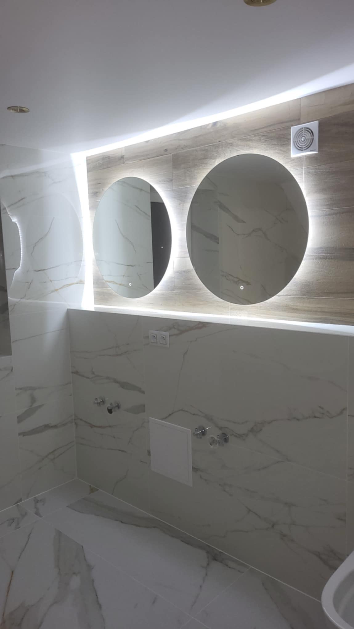 Wykończona łazienka z marmurowymi płytkami, dwoma okrągłymi lustrami z podświetleniem LED i wentylatorem na ścianie, prezentująca nowoczesny design.