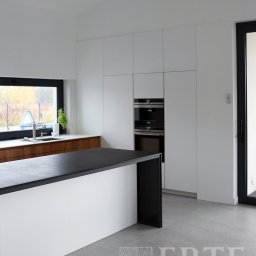 Erte Rafał Tałady Stolarstwo Meblowe - Minimalistyczna kuchnia z wyspą i zabudową szafek w kolorze białym, czarny blat wyspy, okno z widokiem na zewnątrz, szara podłoga, nowoczesne piekarniki w zabudowie.