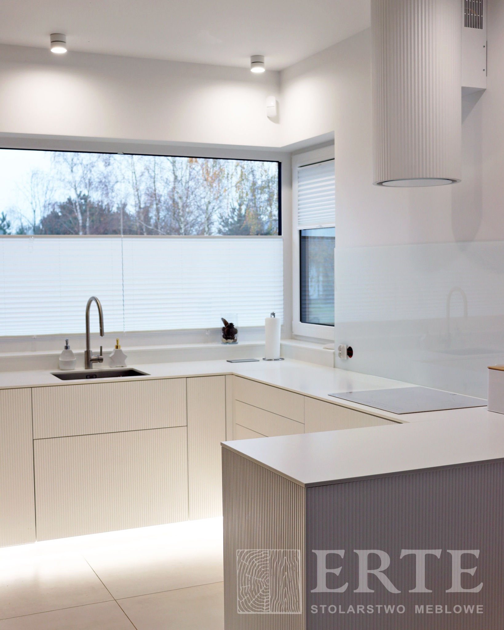 Jasna, minimalistyczna kuchnia z ryflowanymi frontami szafek, dużym oknem z widokiem na drzewa i wyspą kuchenną z logo firmy.