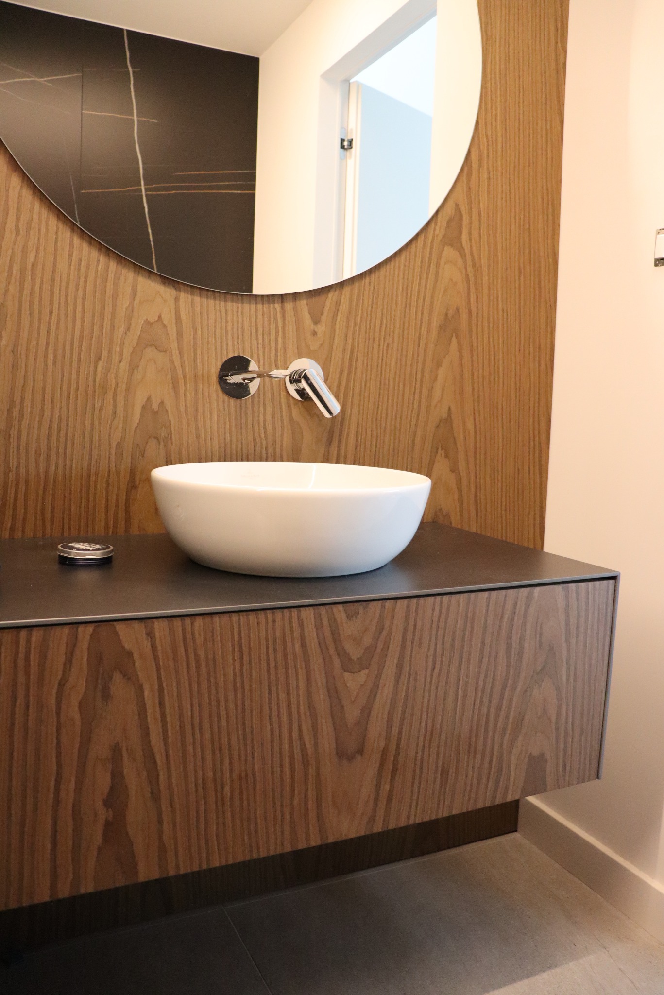 Nowoczesna łazienka z umywalką nablatową, drewnianą szafką pod umywalką i okrągłym lustrem z czarnym akcentem na ścianie.