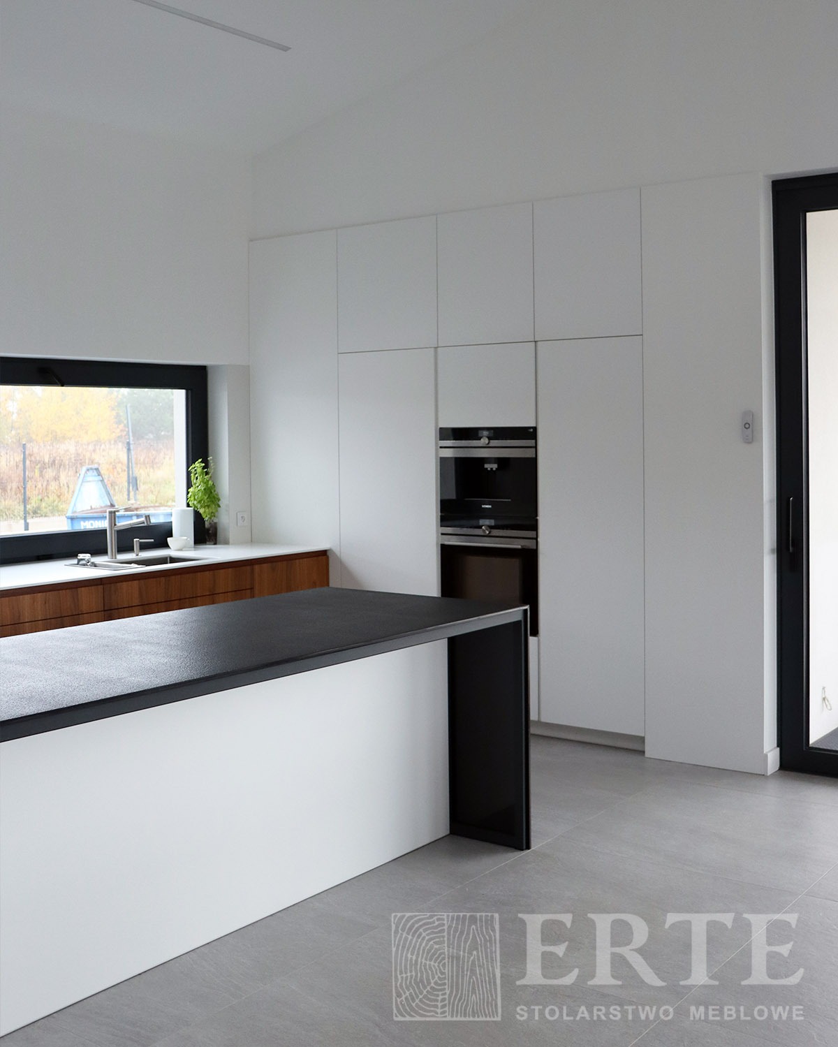Minimalistyczna kuchnia z wyspą i zabudową szafek w kolorze białym, czarny blat wyspy, okno z widokiem na zewnątrz, szara podłoga, nowoczesne piekarniki w zabudowie.