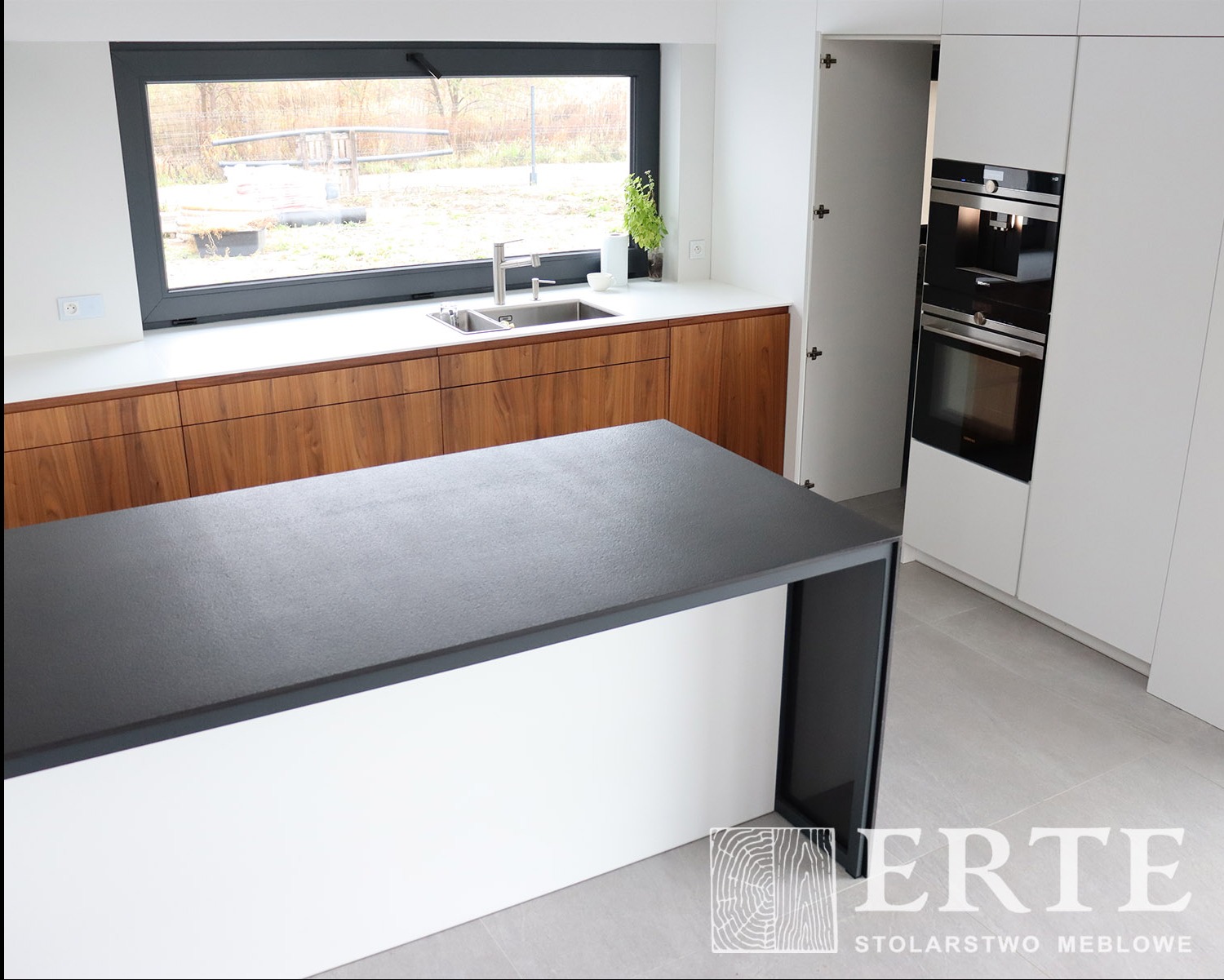 Nowoczesna kuchnia z wyspą: ciemny blat wyspy kontrastuje z białymi frontami i drewnianymi szafkami pod oknem. Widoczny zlew, bateria i wbudowane sprzęty AGD. Otwarta szafka po prawej stronie.