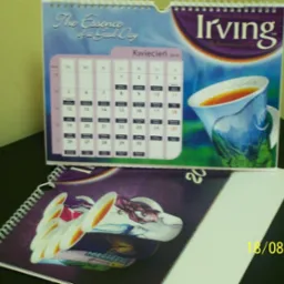 Dwa kalendarze spiralne, jeden stojący z motywem herbaty Irving i widocznym kwietniem 2010, drugi leżący z podobną grafiką filiżanki, na ciemnym blacie.