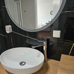 Shany up - Nowoczesna łazienka z okrągłym lustrem, białą umywalką nablatową, chromowaną baterią i czarnymi płytkami z złotymi żyłkami.