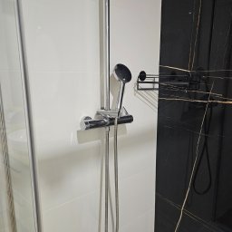 Shany up - Nowoczesna kabina prysznicowa z chromowaną armaturą na tle białych płytek i czarnego marmuru z złotymi żyłami, widoczna półka na kosmetyki.