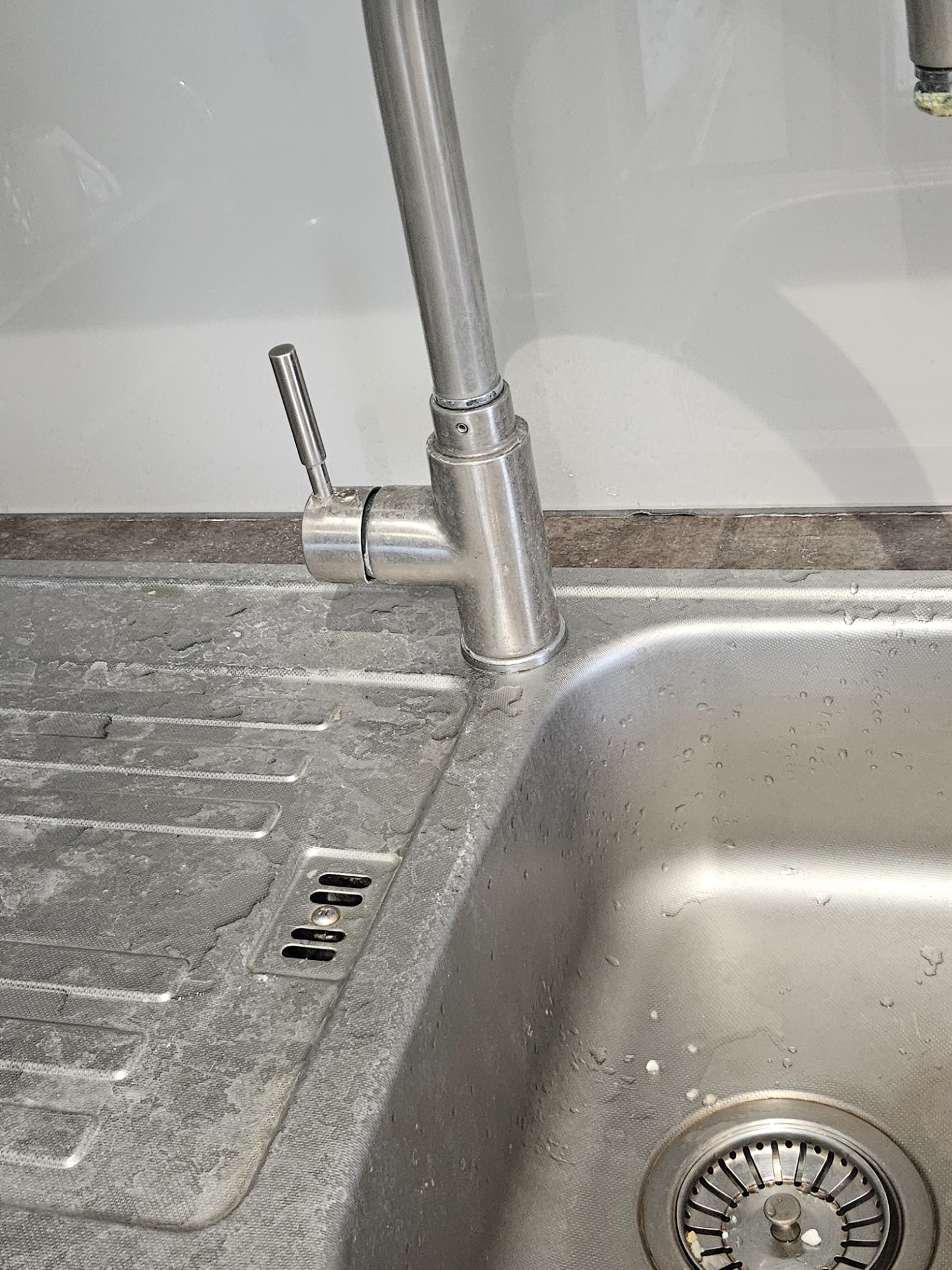 Zanieczyszczony, metalowy zlew kuchenny z osadem i kroplami wody, widoczna bateria z pojedynczą dźwignią i odpływ z resztkami.