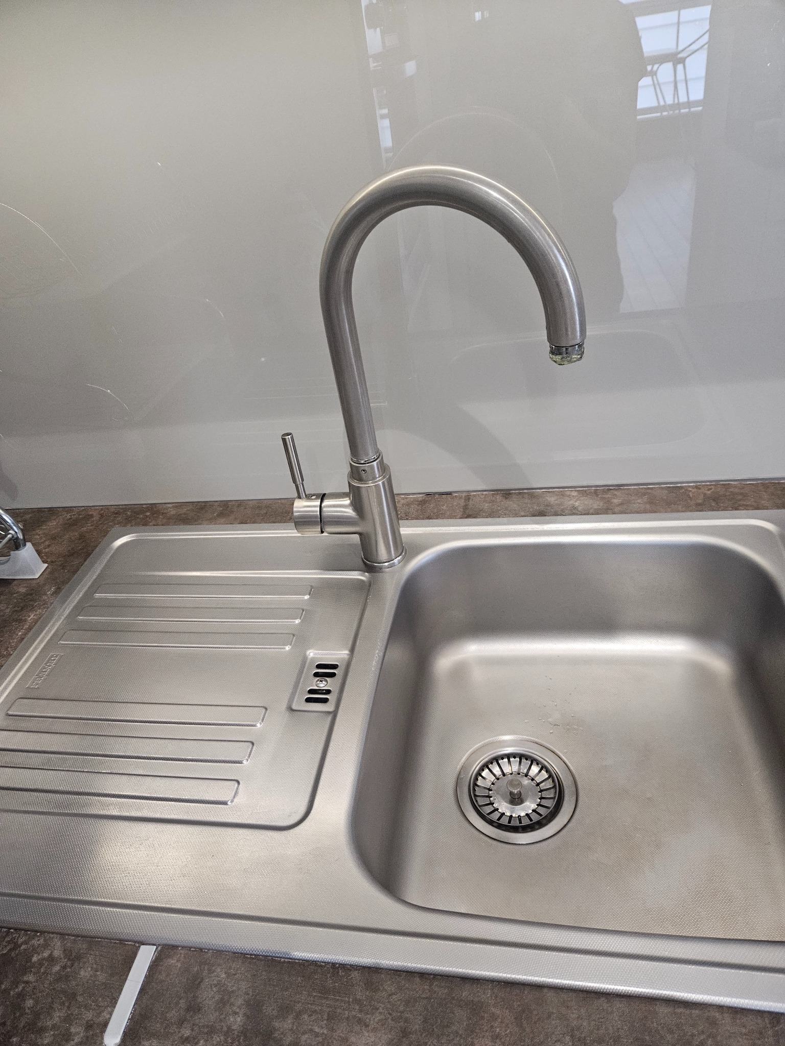 Błyszczący, metalowy zlew jednokomorowy z ociekaczem i wysoką, łukowatą baterią, na tle jasnoszarej ściany i blatu.