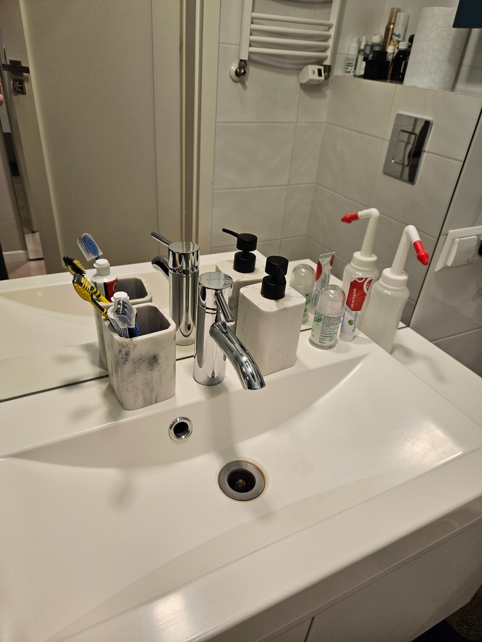 Umywalka z białą armaturą, dozownikami mydła, szczoteczkami do zębów i kosmetykami na blacie, odbicie w lustrze.