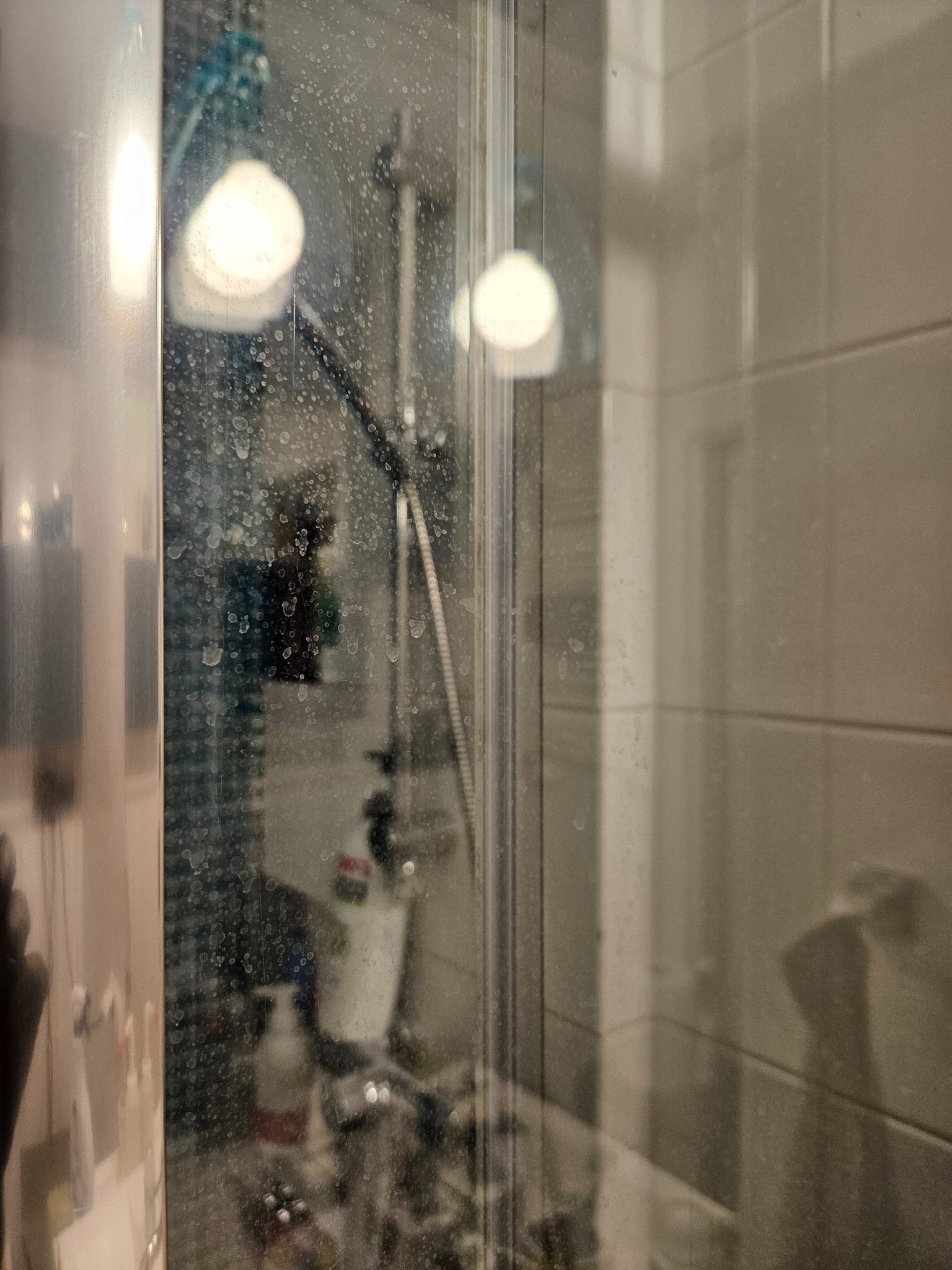 Zabrudzona kabina prysznicowa z widocznymi zaciekami i osadem z mydła na szklanych drzwiach, armatura prysznicowa i butelki kosmetyków w tle, płytki ceramiczne na ścianie.