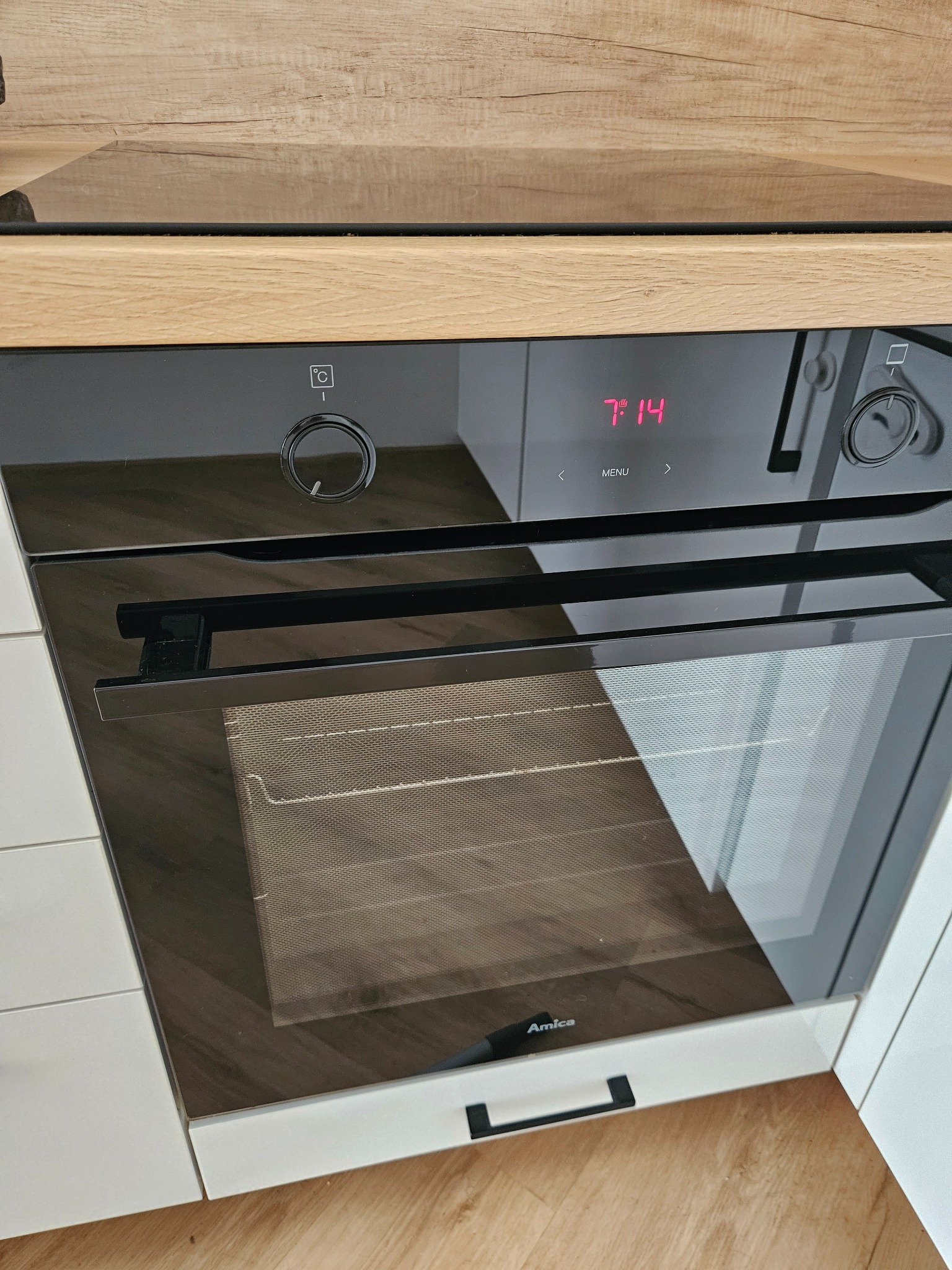 Wbudowany piekarnik Amica w nowoczesnej kuchni, z wyświetlaczem wskazującym godzinę 7:14, umieszczony w zabudowie z drewnianym blatem i białymi szafkami.
