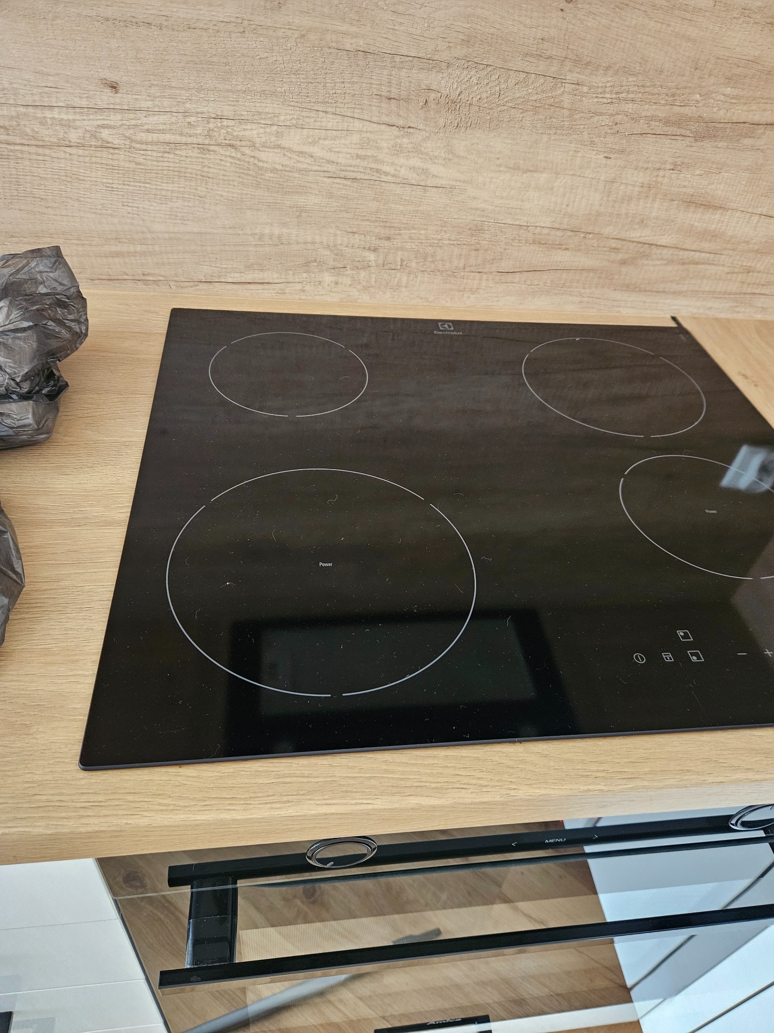 Czarna płyta indukcyjna Electrolux z czterema polami grzewczymi, zamontowana w blacie o jasnym, drewnopodobnym wzorze, z widocznym fragmentem piekarnika w dolnej części kadru i zwiniętym workiem...