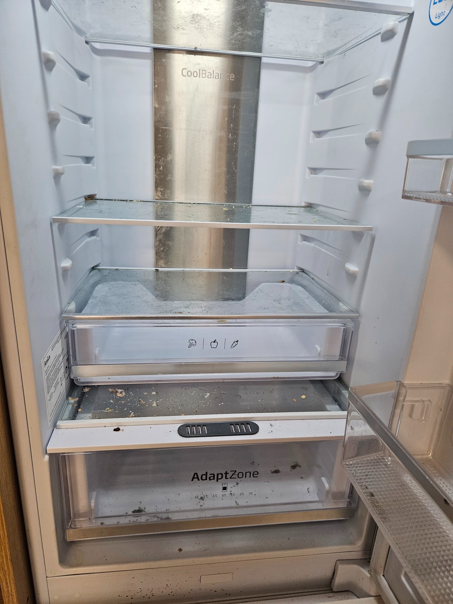 Wnętrze brudnej lodówki marki Amica z półkami i szufladami pokrytymi okruchami jedzenia i plamami.