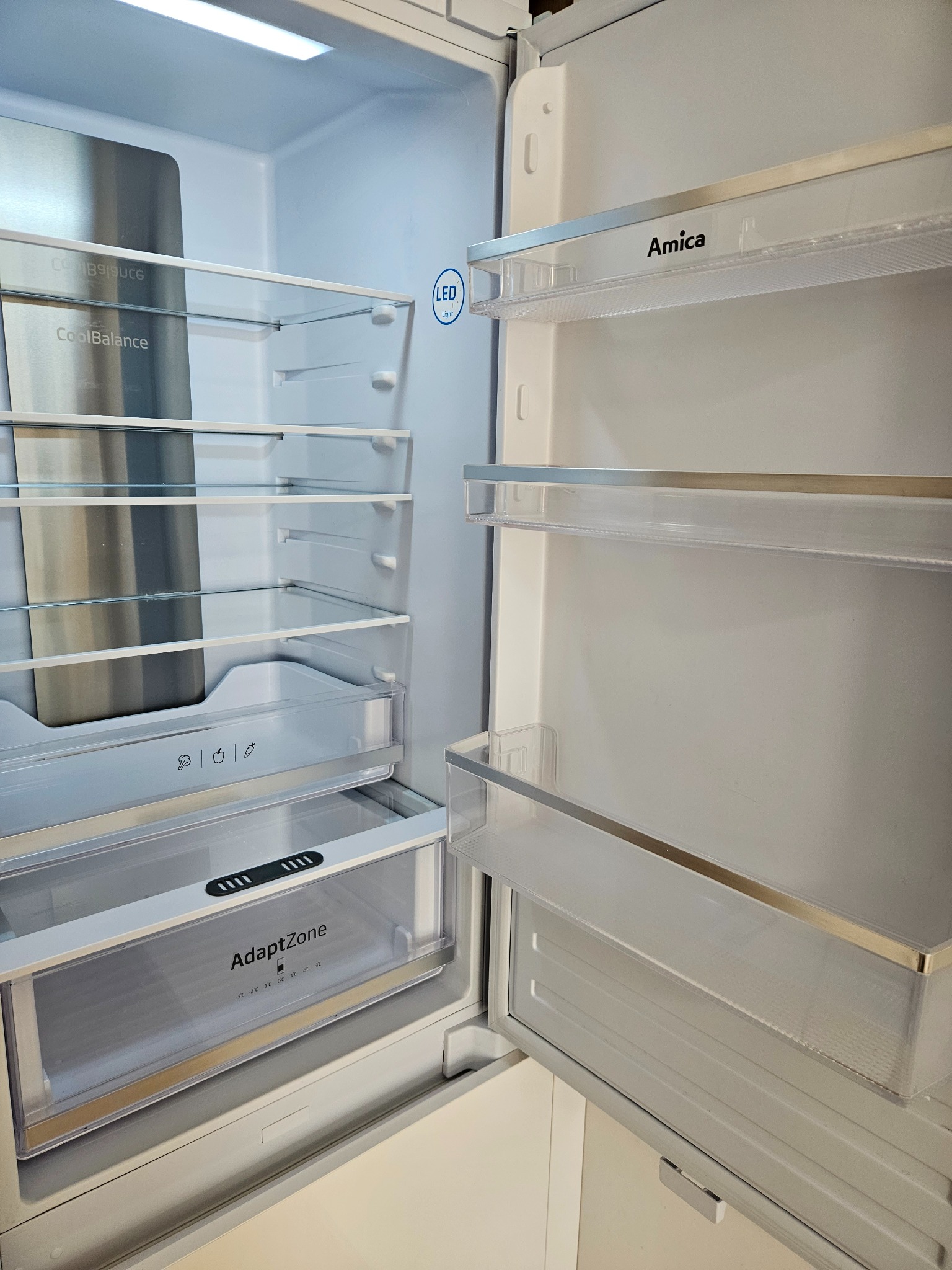 Wnętrze otwartej, pustej lodówki marki Amica z półkami z przezroczystego tworzywa, oświetleniem LED i specjalną szufladą AdaptZone na dole.