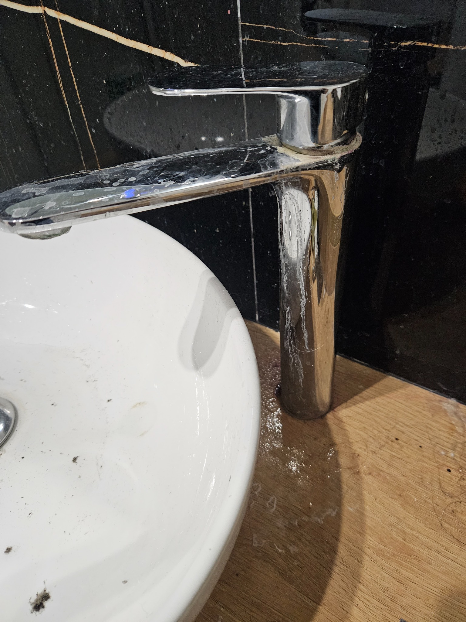 Zanieczyszczona biała umywalka z chromowaną baterią pokrytą zaciekami, na tle czarnych płytek z marmurowym wzorem i drewnianego blatu.