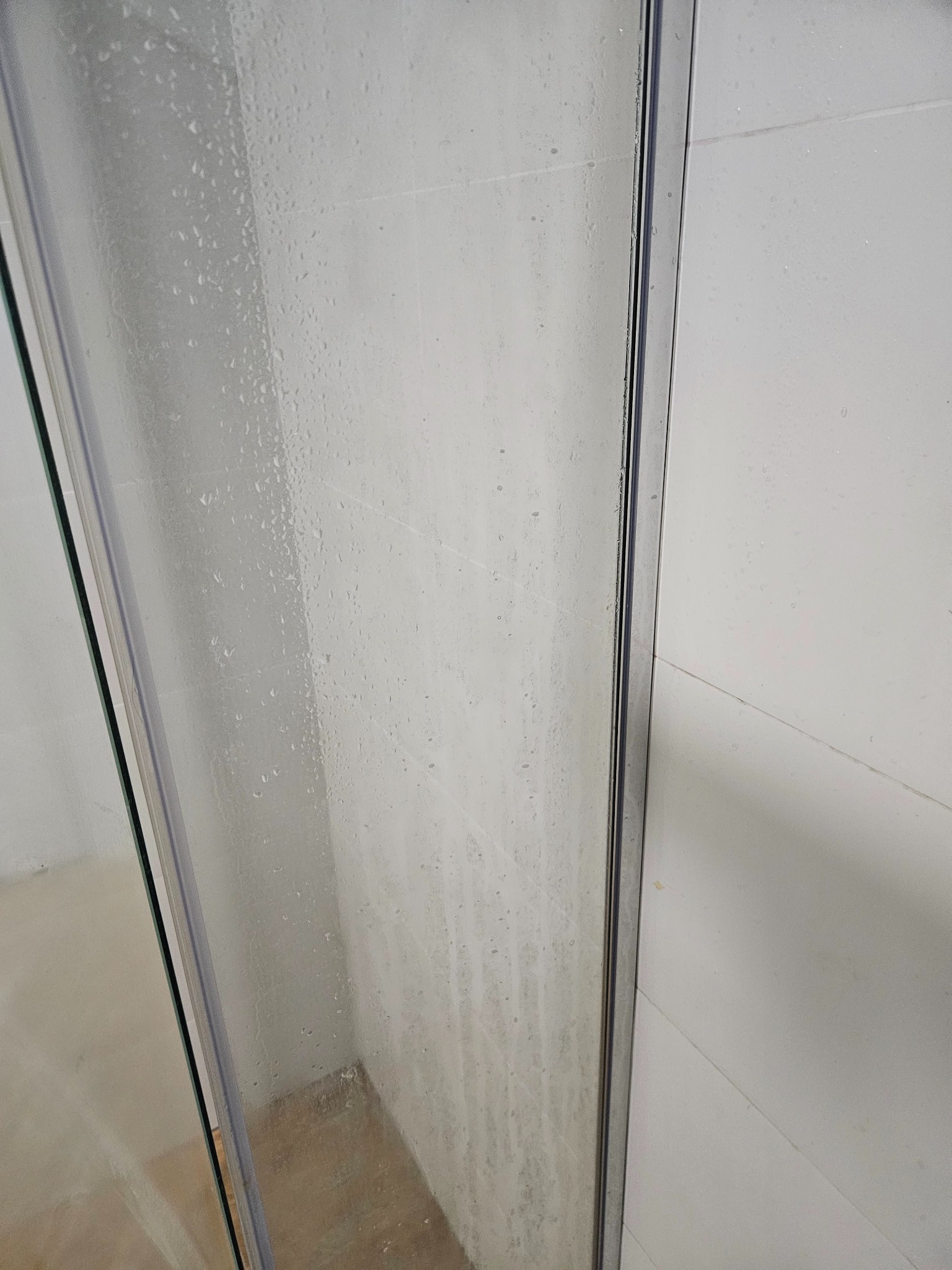 Szklana kabina prysznicowa z widocznymi zaciekami i osadami z mydła na szkle, przylegająca do białych, matowych płytek ściennych.