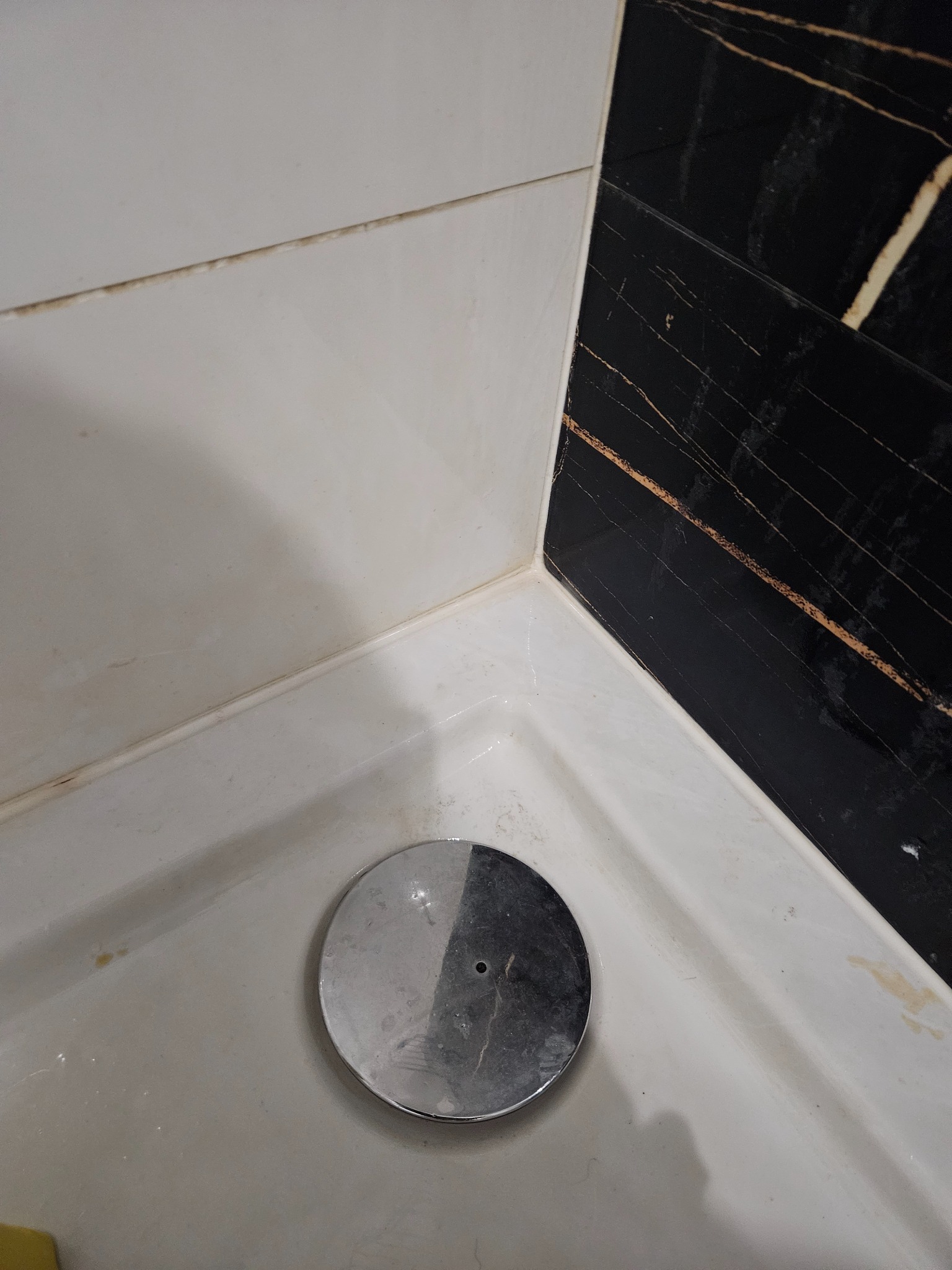 Niedoczyszczony odpływ brodzika z chromowaną pokrywką, widoczne zacieki i osady na białym brodziku i czarnych marmurowych płytkach ściennych.