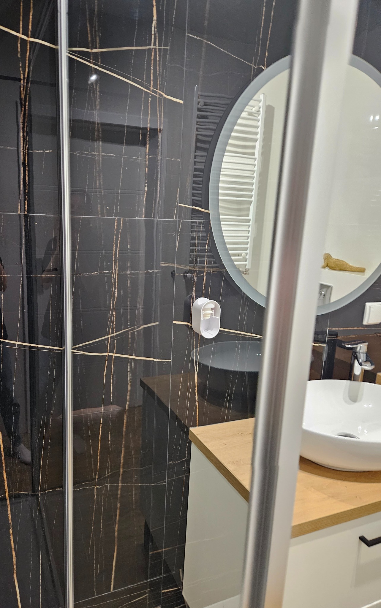 Nowoczesna łazienka z czarnymi płytkami imitującymi marmur ze złotymi żyłami, kabiną prysznicową z przezroczystego szkła, okrągłym lustrem i białą umywalką na drewnianym blacie.