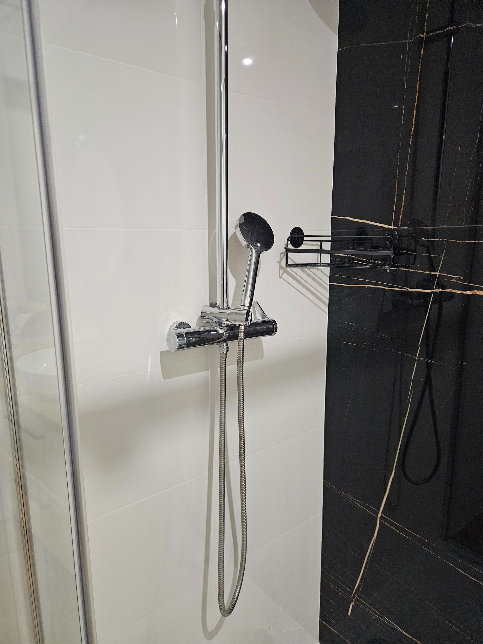 Nowoczesna kabina prysznicowa z chromowaną armaturą na tle białych płytek i czarnego marmuru z złotymi żyłami, widoczna półka na kosmetyki.