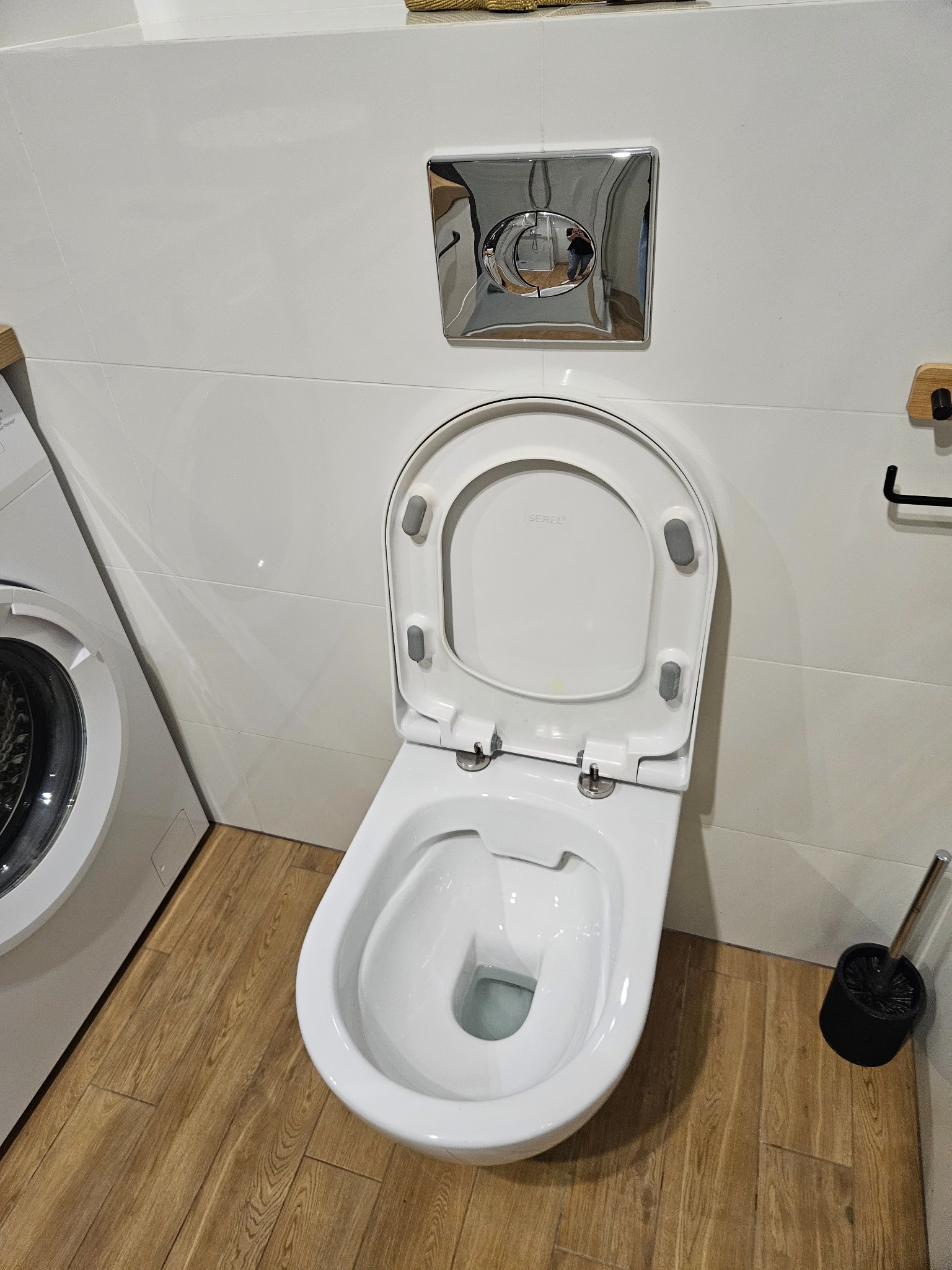 Biała toaleta podwieszana z otwartą deską, chromowany przycisk spłuczki, drewnopodobne płytki podłogowe, pralka w tle, szczotka do WC obok muszli.